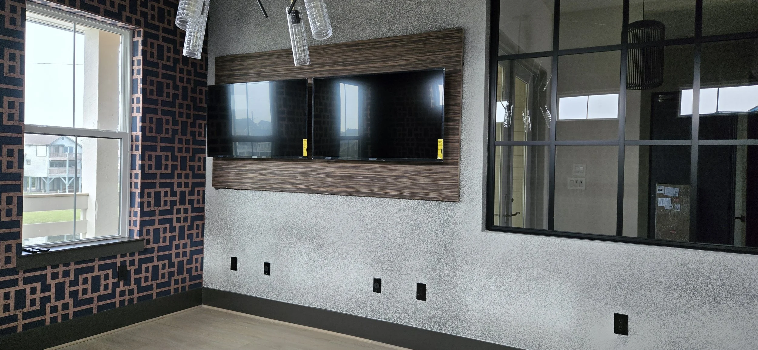 Two wall-mounted TVs set into a wood-paneled recess on a textured wall beside a large black-framed window, during construction