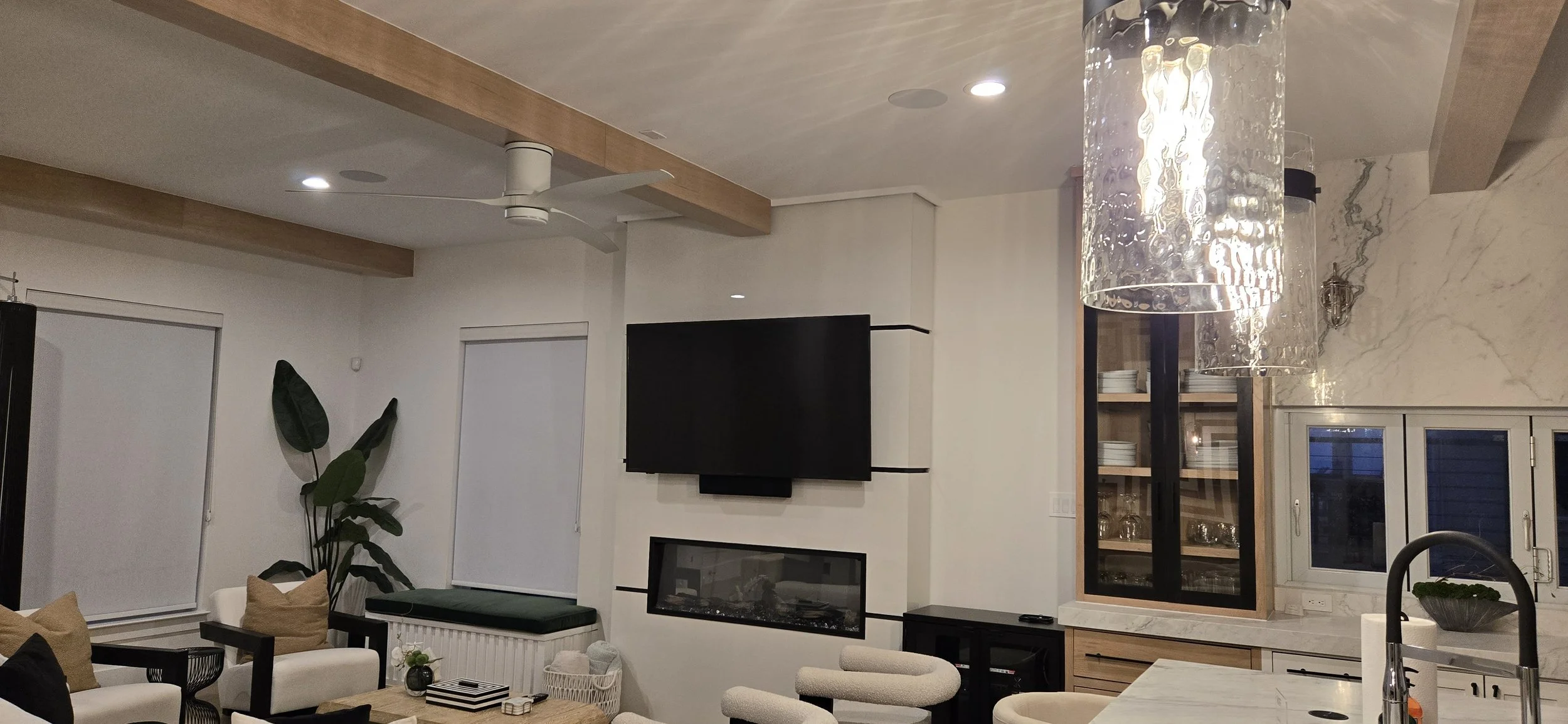 Open living room with a wall-mounted TV above a linear fireplace, white walls, wood ceiling beams, and a glass pendant light