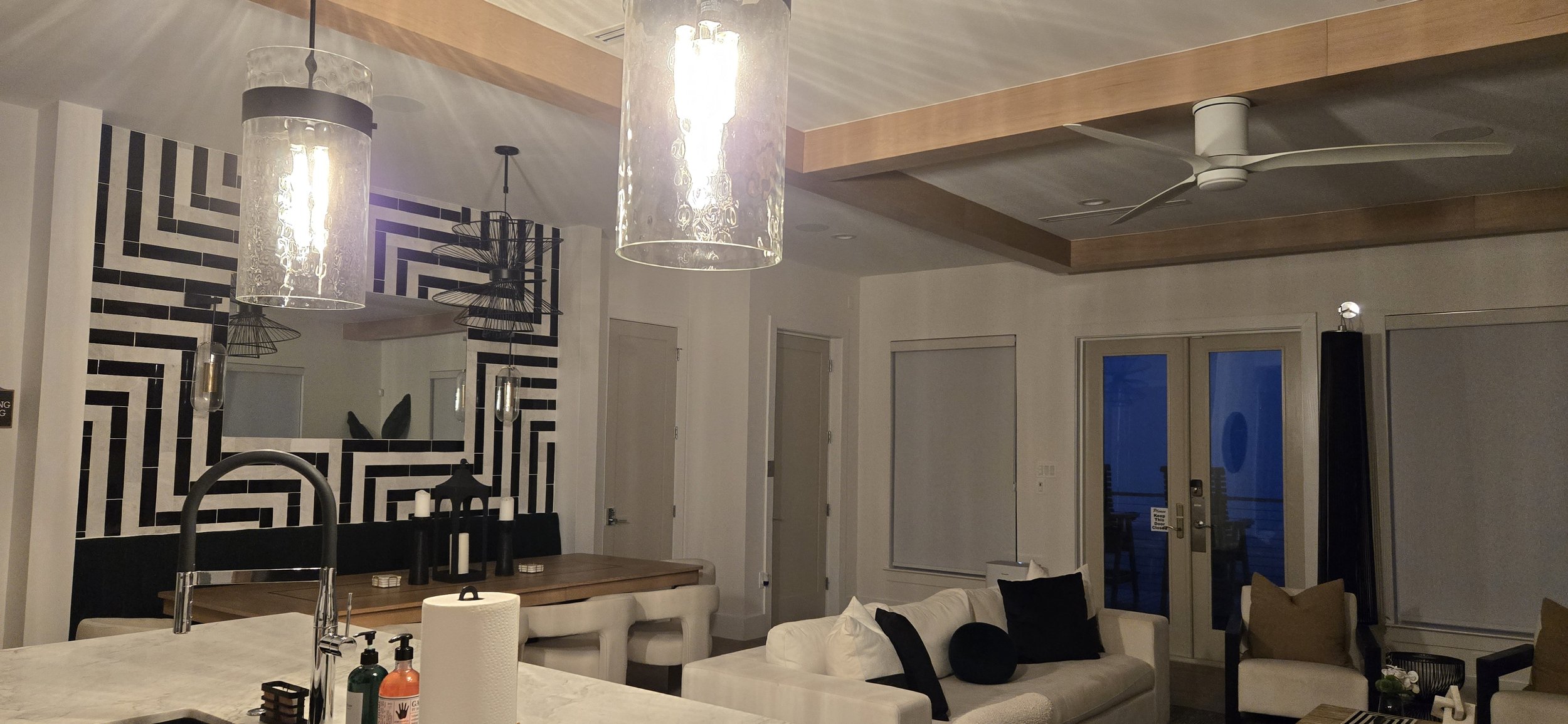 Open kitchen, dining, and living area with a black-and-white geometric accent wall, glass pendant lights, and wood ceiling beams