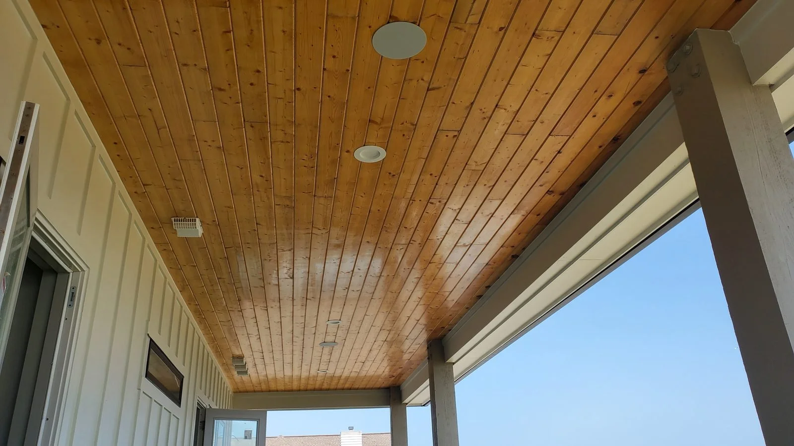 Weatherproof speakers recessed in the stained wood plank ceiling of a covered porch, with a board-and-batten wall and blue sky beyond.