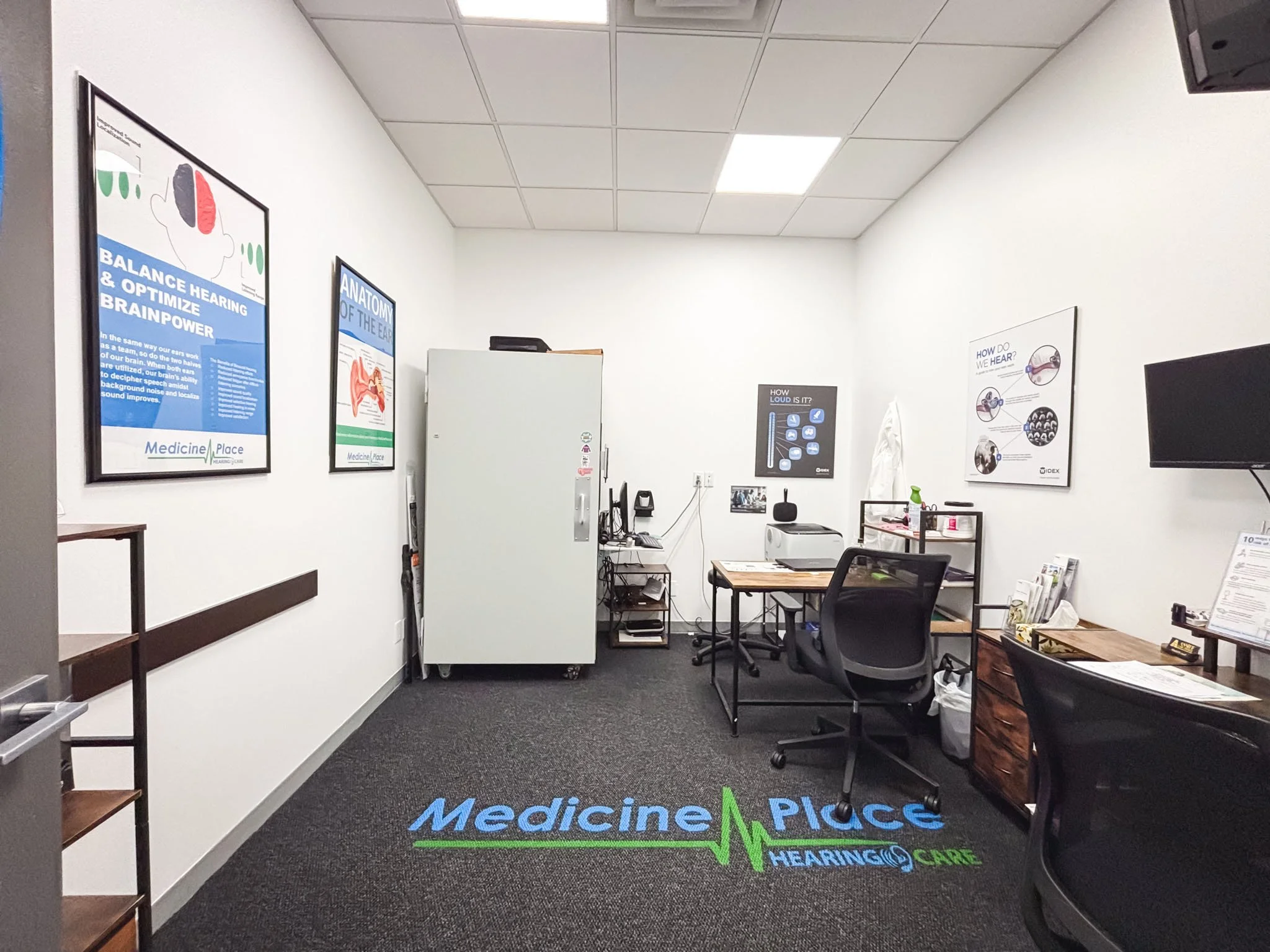 An office room with white walls and a dark carpeted floor. There are three framed posters on the left wall related to hearing and ear anatomy. In the center, there is a gray storage cabinet, a small desk with a computer, and a black office chair. A laminated floor mat with the words "Medicine Place Hearing Care" is in the foreground. On the right side, there is a laminating poster on the wall, a small cart with medical supplies, and another desk with papers and chairs.