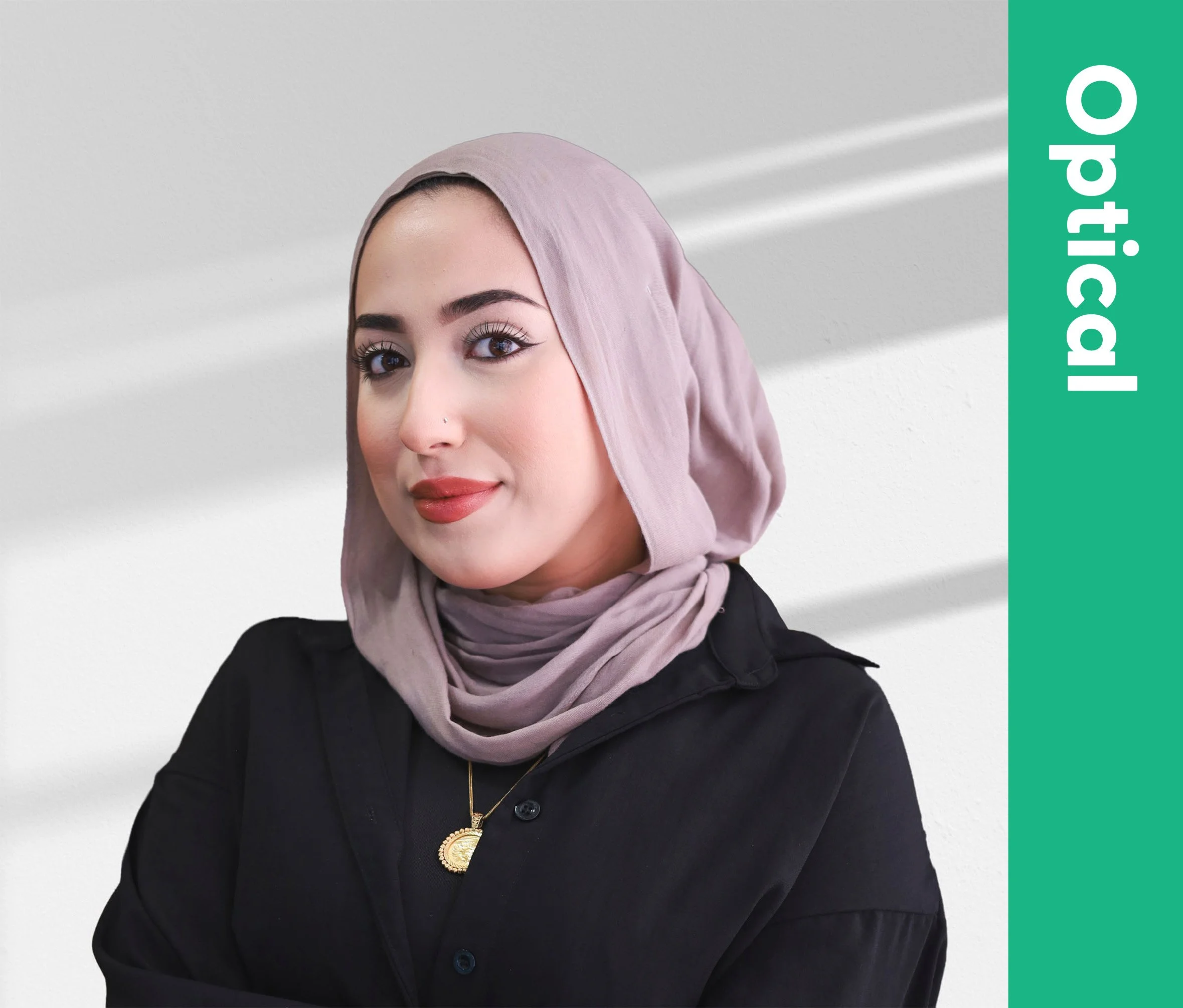 A woman with dark eyes and eyebrows, wearing a light pink hijab and a black shirt with a gold necklace, smiling softly against a plain light background.