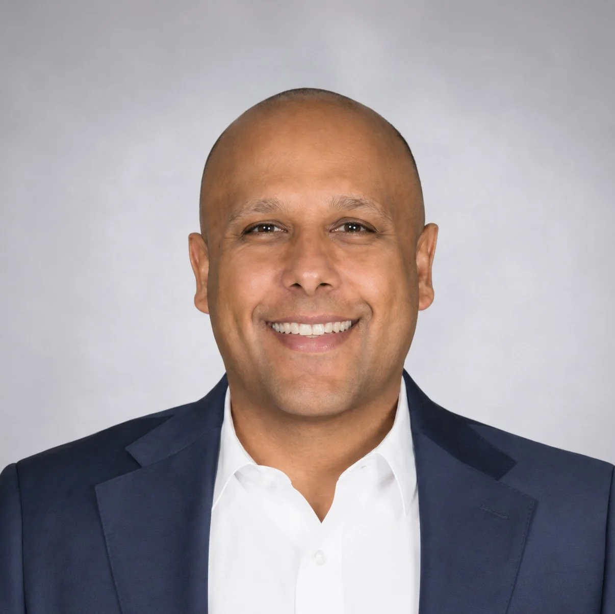 A smiling man with a shaved head wearing a navy blazer and white shirt against a neutral background.