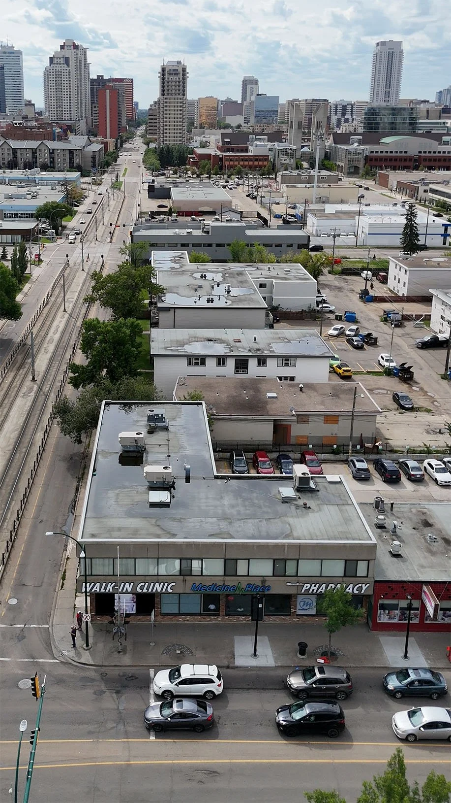 Cityscape view from an elevated position showing a mix of commercial and residential buildings, including a healthcare clinic on the ground floor, parking lots, and streets with cars, with a skyline of tall buildings in the background under a cloudy 