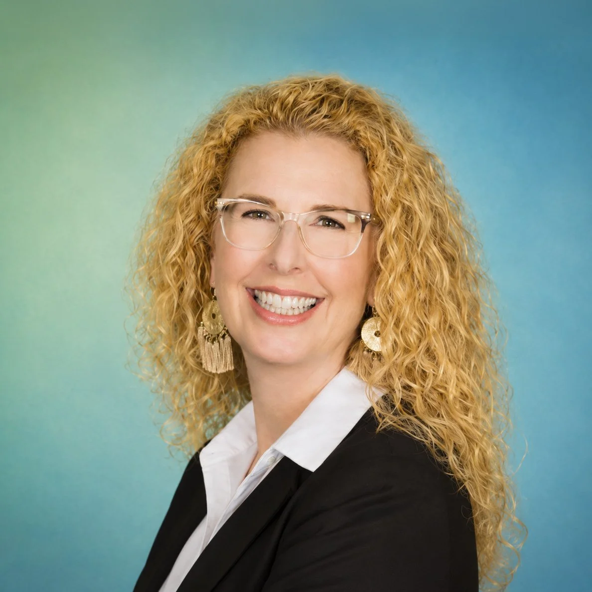 A woman with curly blonde hair, wearing glasses, large earrings, a white collared shirt, and a black blazer, smiling against a gradient blue and green background.