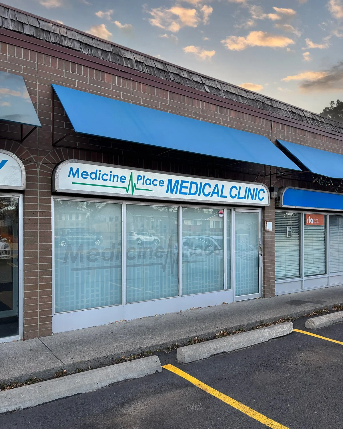 Front view of Medicine Place Medical Clinic with blue awnings and glass windows, parking lot in front, cloudy sky overhead.