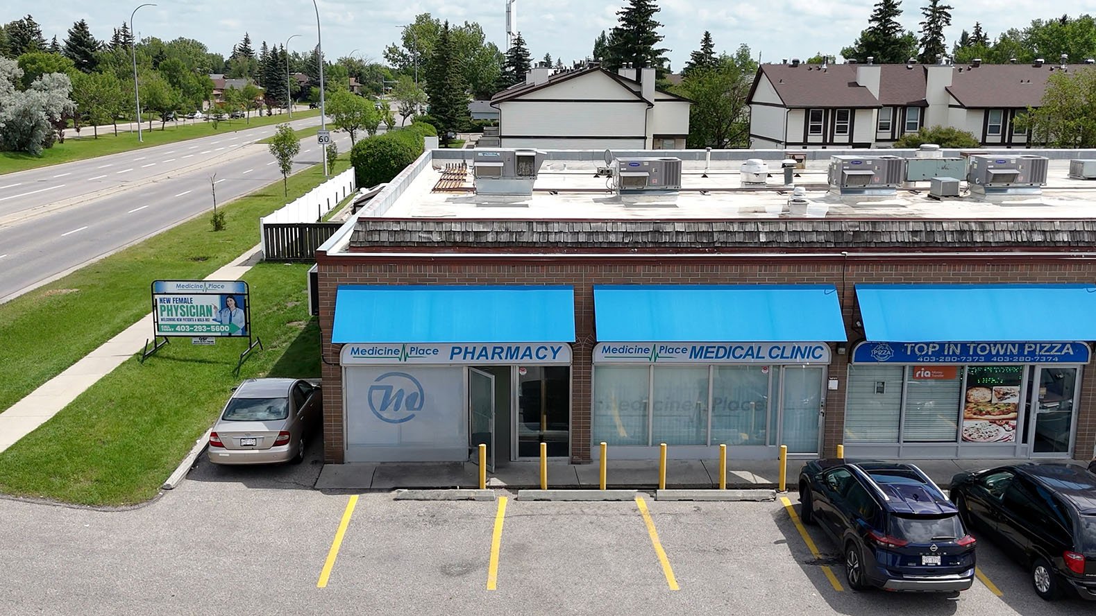 View of a building with three storefronts: a pharmacy, a medical clinic, and a pizza restaurant with blue awnings and parking spaces in front. There is a sign outside advertising a female physician and a highway with trees and houses in the backgroun
