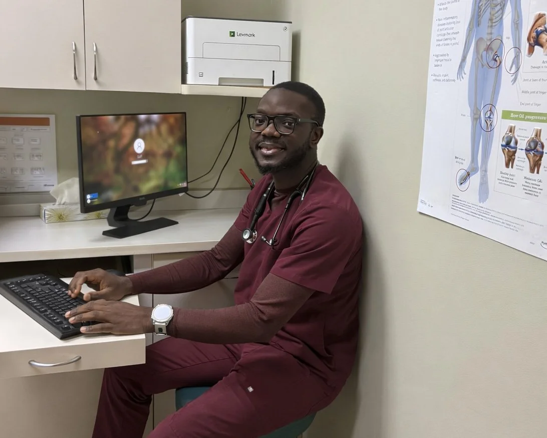 A male healthcare professional sitting at a desk in a medical office, wearing maroon scrubs, glasses, with a stethoscope around his neck, smiling at the camera.