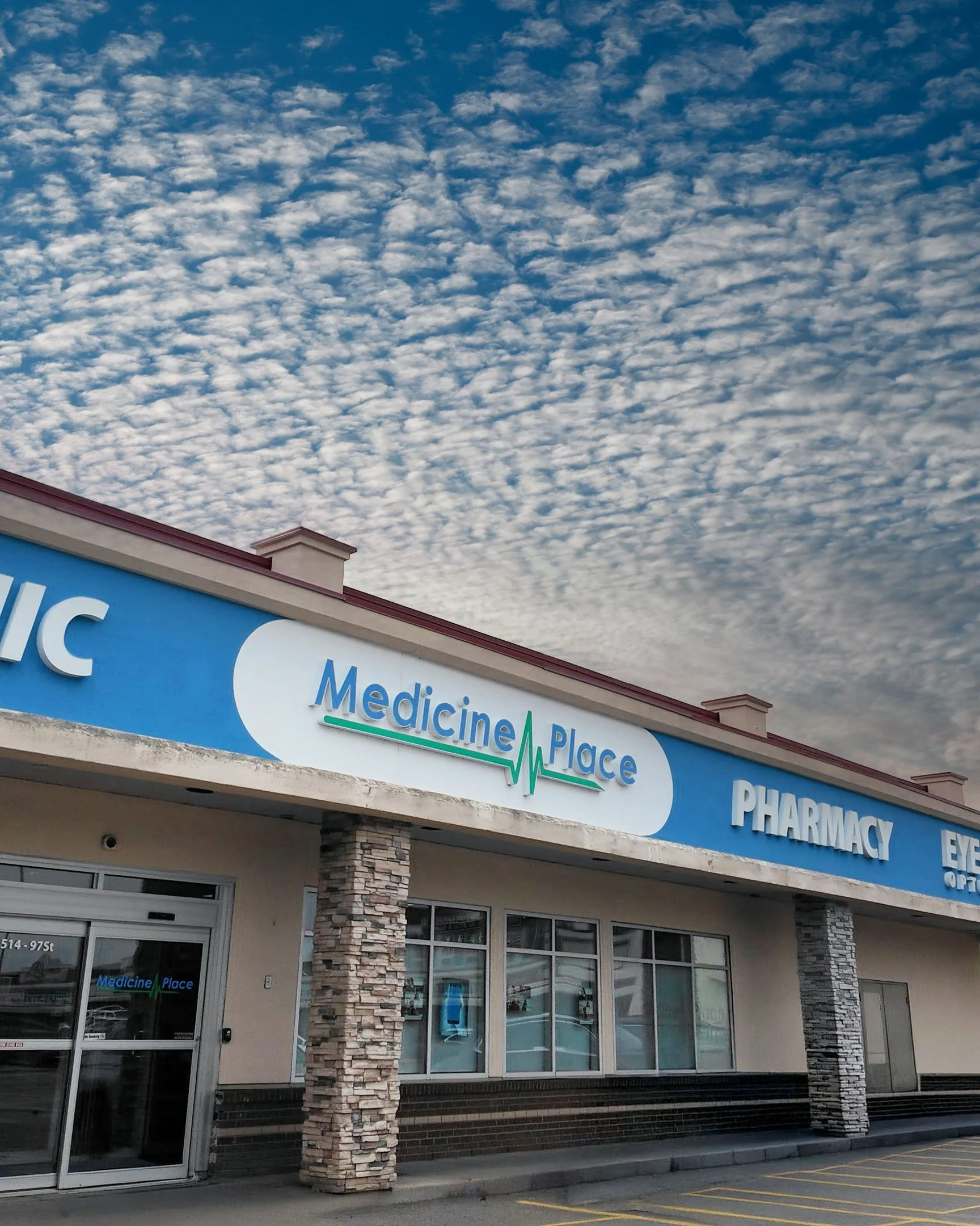 Front view of Medicine Place, a pharmacy/store with a blue sign, stone columns, glass windows, and a cloudy sky above.