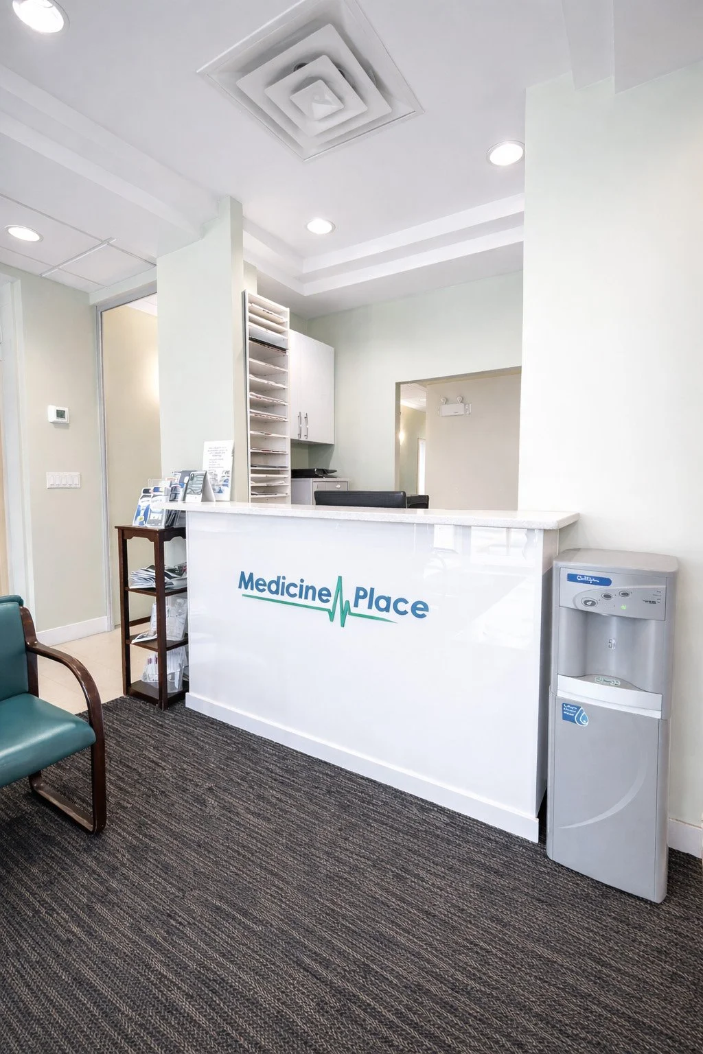 Reception area at a medical office with a white counter signed 'Medicine Place,' a water dispenser on the right, green waiting chairs on the left, and light-colored walls with ceiling lights.