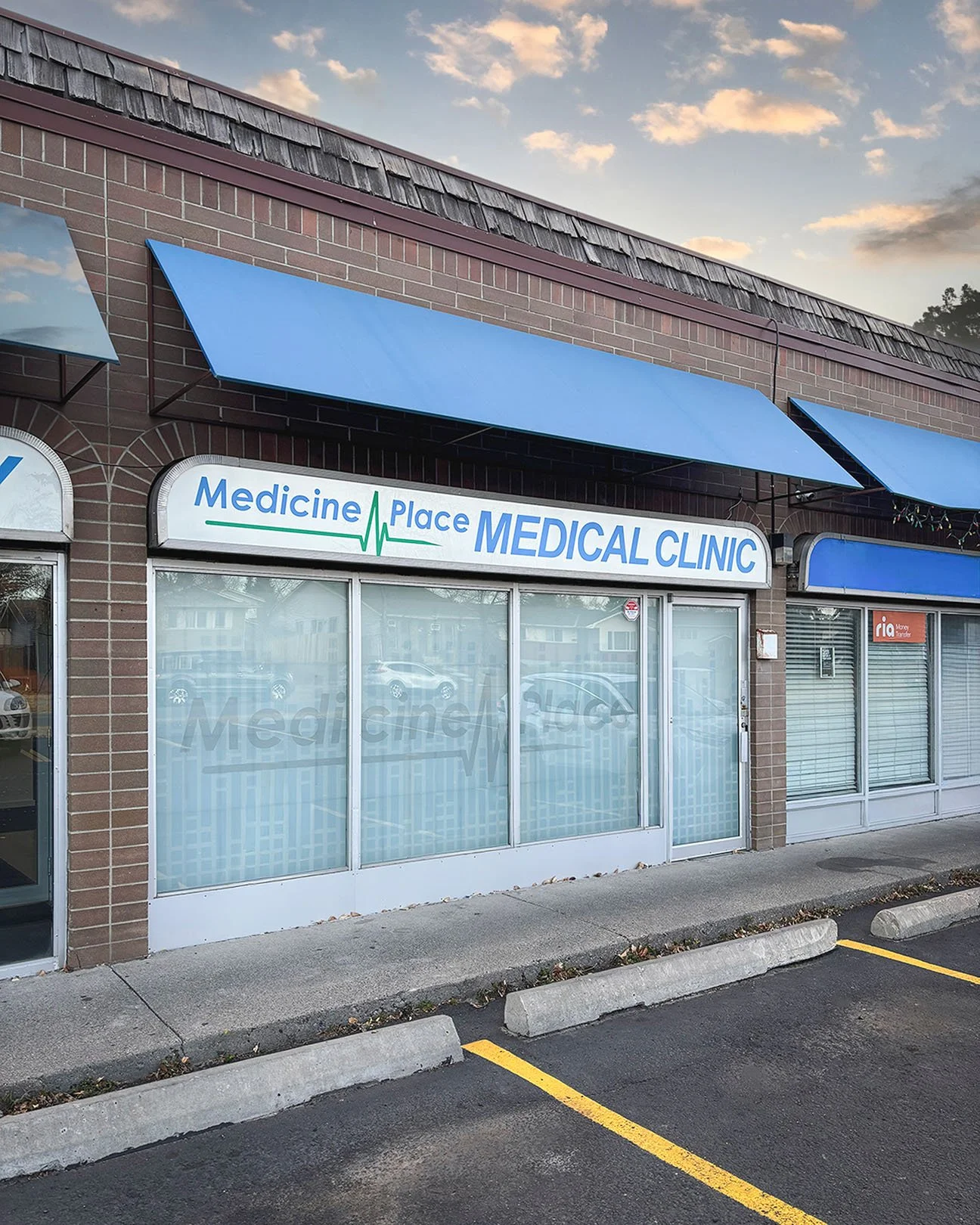 Exterior view of a medical clinic called Medicine Place Medical Clinic with blue awnings, brick facade, large windows, and parking lot in front.
