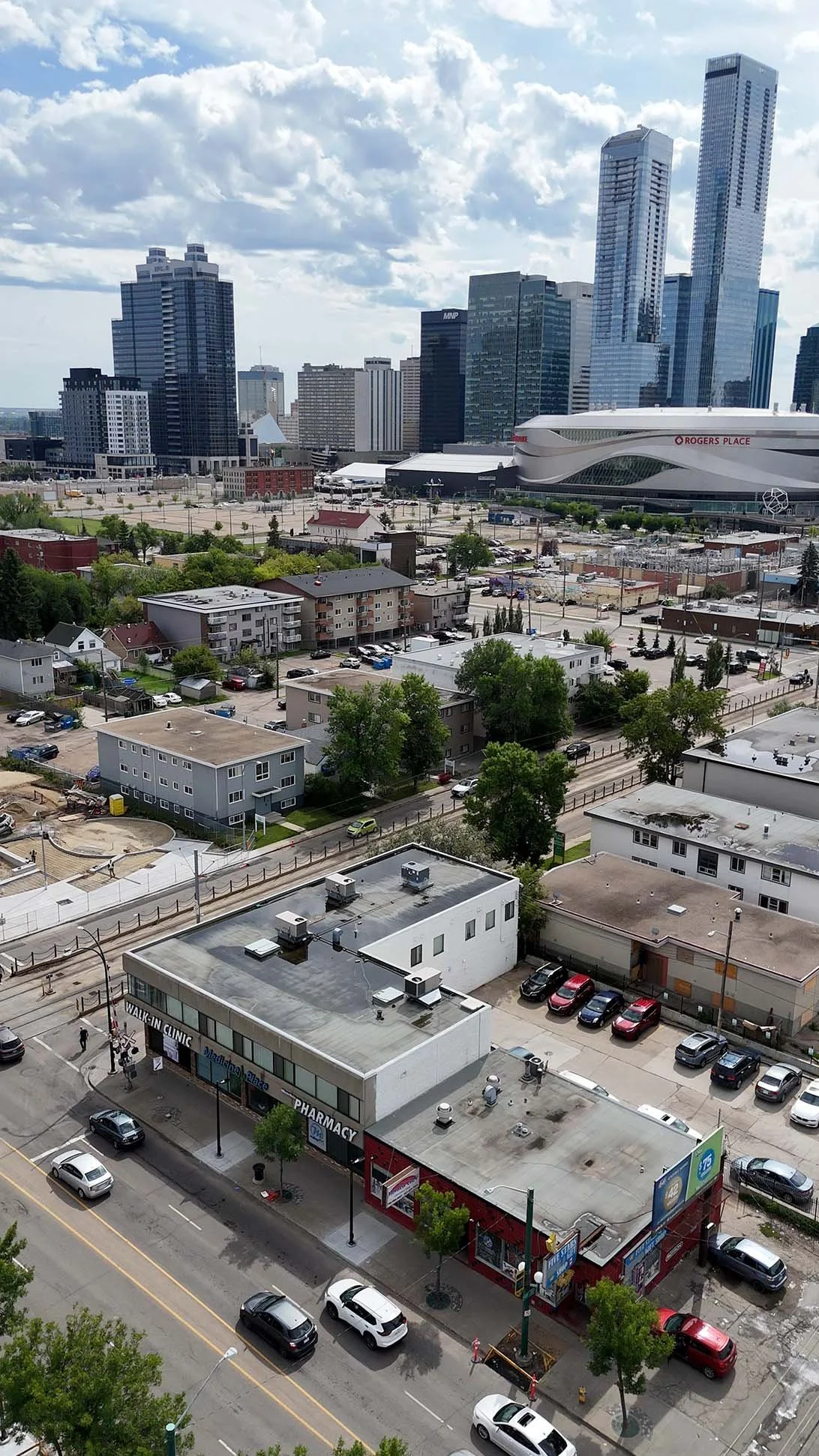 A cityscape showing a mix of residential and commercial buildings with skyscrapers in the background, including Rogers Place arena. The foreground includes a pharmacy building, parking lots, and streets with cars, trees, and pedestrians.