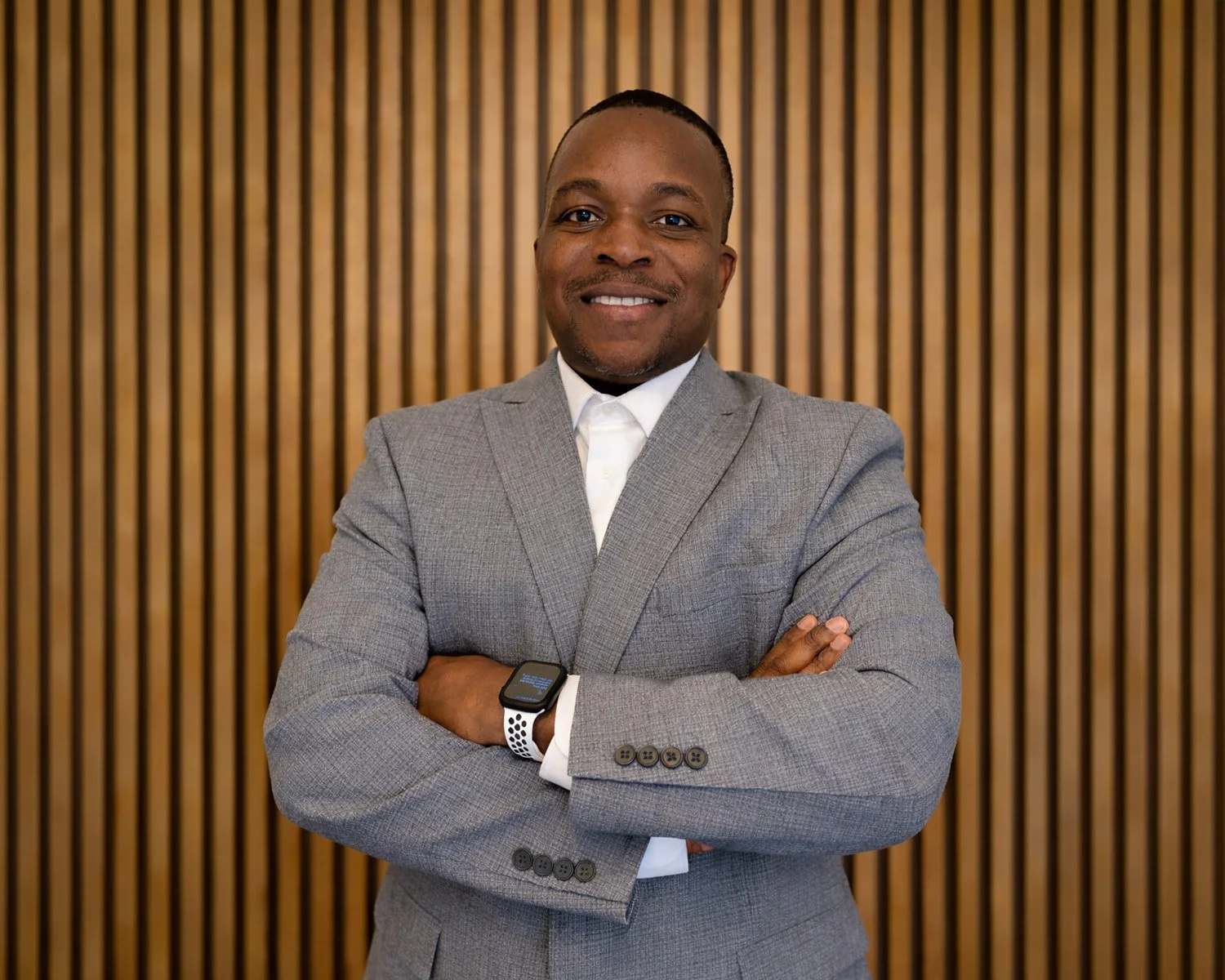 A man in a gray suit with crossed arms, smiling, standing in front of a wooden slat wall.