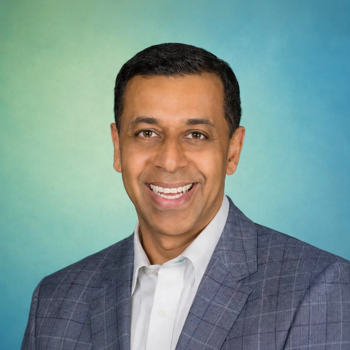 Headshot of a smiling man wearing a navy checked blazer and white shirt, against a blue and green gradient background.