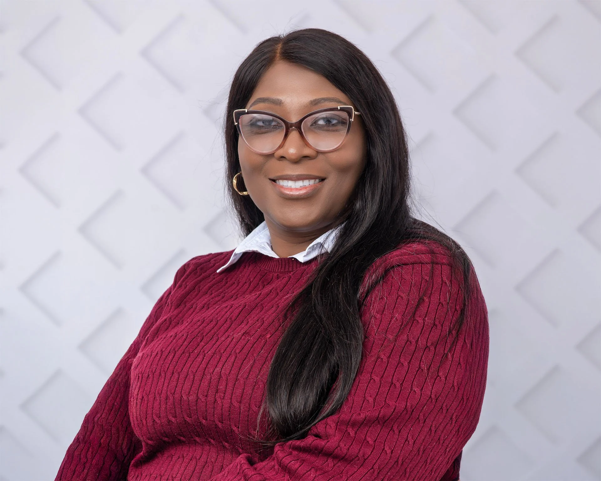 Portrait of a woman with long dark hair, wearing glasses, a white shirt, and a red sweater, smiling at the camera with a textured white wall background.
