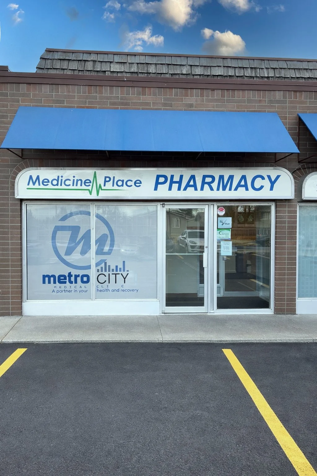Front view of a pharmacy store named Medicine Place Pharmacy with blue awning, large glass window, and parking lot with yellow lines.