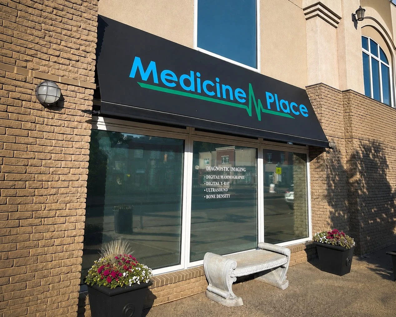 Exterior view of a medical clinic called 'Medicine Place' with a black awning, large window, flowers in planters, and a white bench outside.