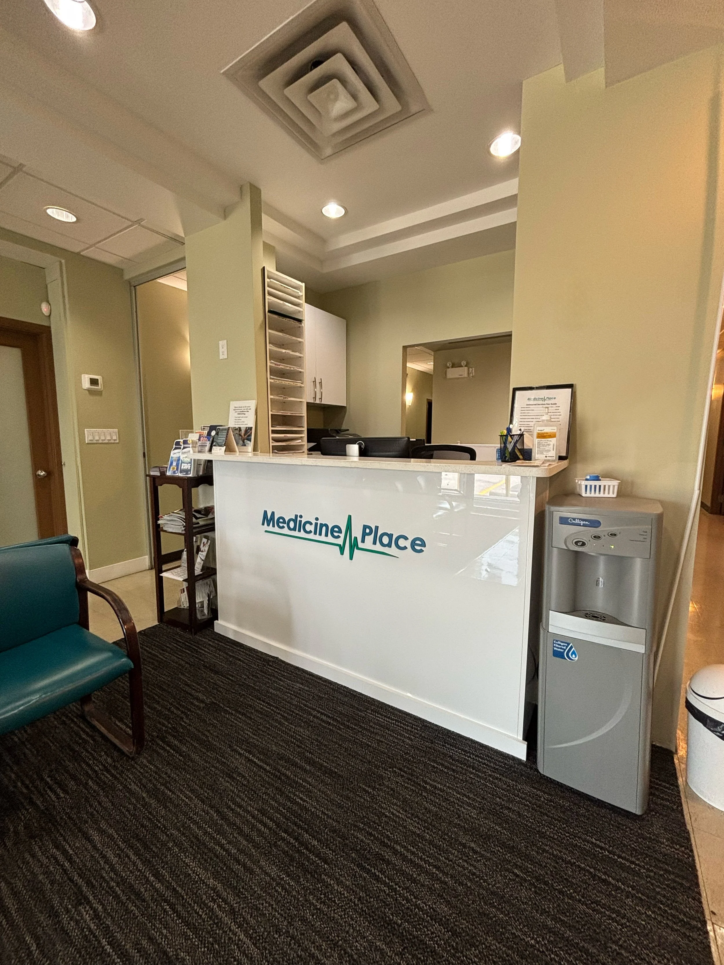 Reception area of Medicine Place clinic with a white reception desk, a water dispenser on the right, a dark green chair to the left, and informational materials on the desk.