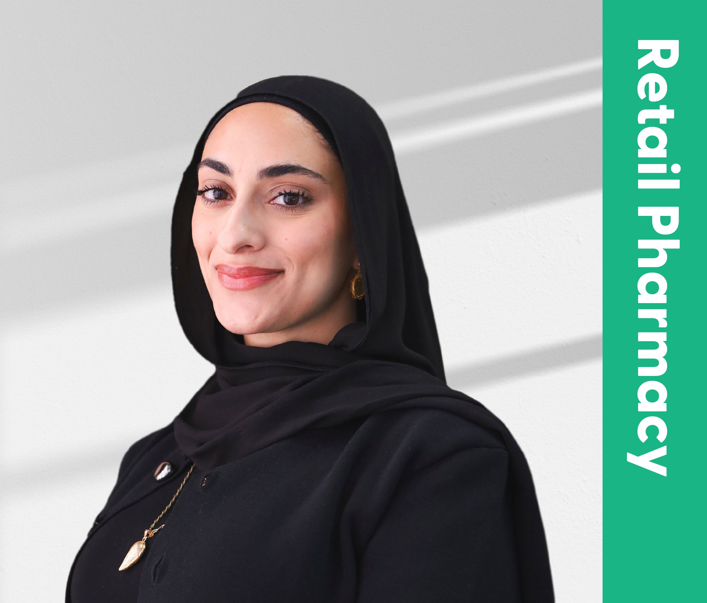 A woman with dark eyes, eyebrows, and hair partially covered by a black headscarf, smiling subtly, wearing a black top or outer garment, against a plain light-colored wall background.