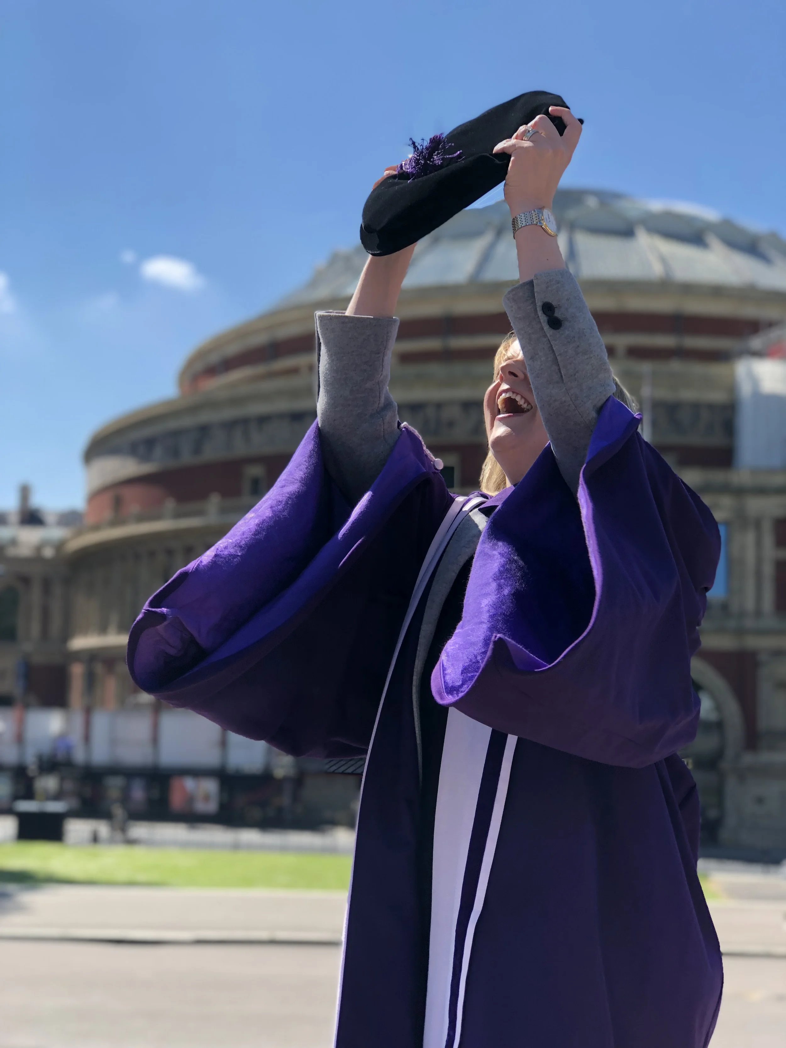Dr Janet Deane in her Imperial College London PhD graduation gown lifting her cap in celebration outside a historic building under a blue sky.
