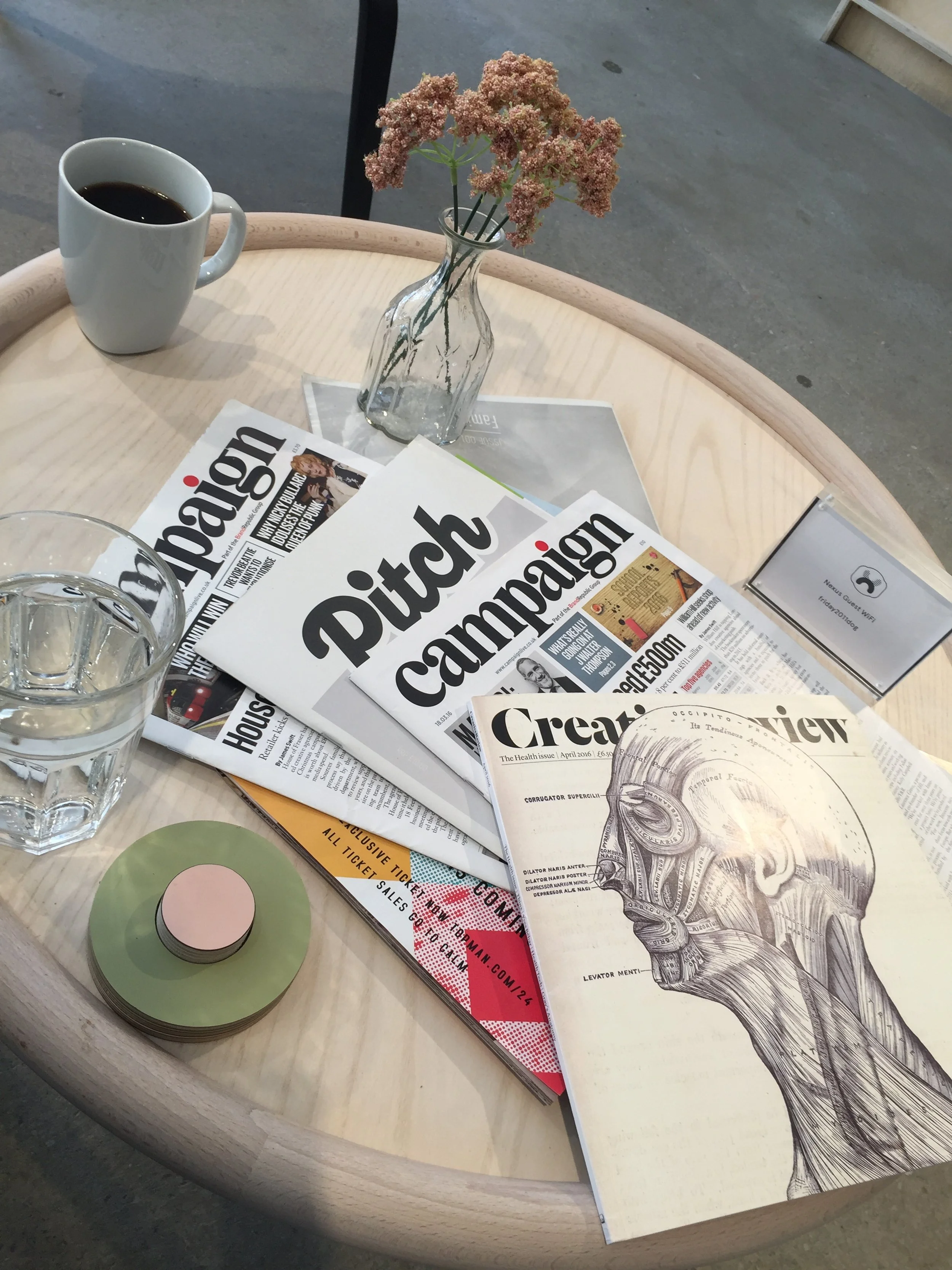 A round wooden table with a glass of water, a white coffee mug filled with coffee, a vase with pink flowers, several magazines including one titled "Pitch Campaign" and "Creative Review," to represent the varied ways in which academics need to work.