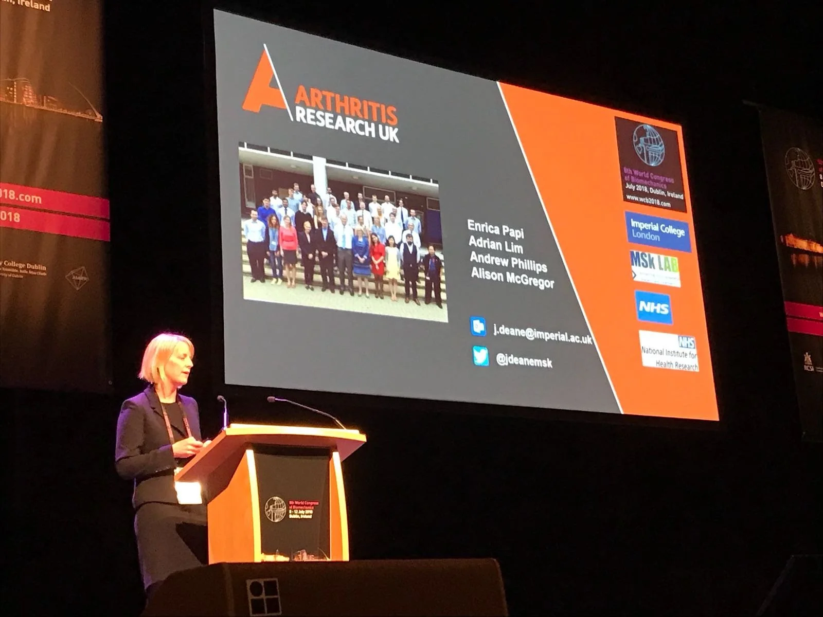 Dr Janet Deane speaking at a podium during a conference with a large screen behind her displaying a presentation slide from her PhD funding body, Versus Arthritis, Formerly Arthritis Research UK.