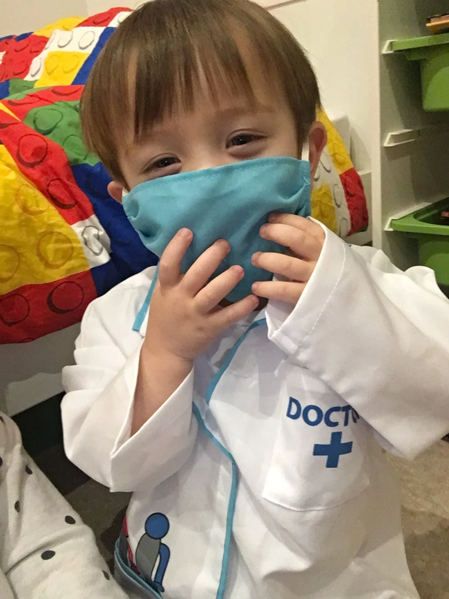 A young child dressed as a doctor wearing a white coat with a blue cross and the word 'DOCTOR' on it, holding a light blue face mask with both hands, smiling to represent the PhD motherhood journey.
