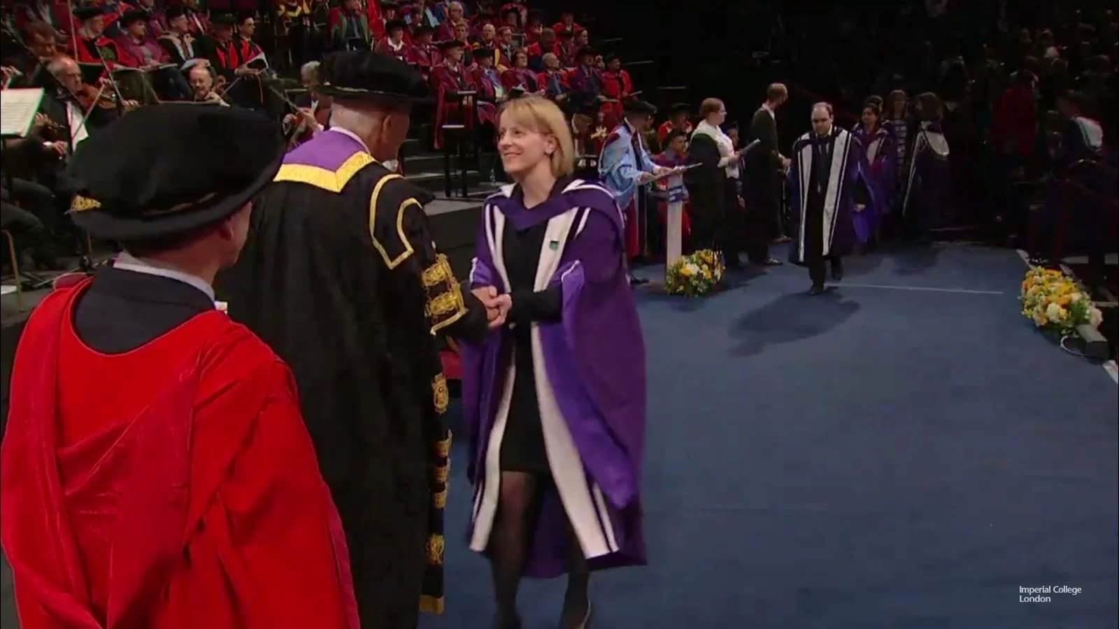 Dr Janet Deane receiving a her PhD from Imperial College London at a graduation ceremony at The Royal Albert Hall, shaking hands with the University President, with other graduates in robes and caps in the background.