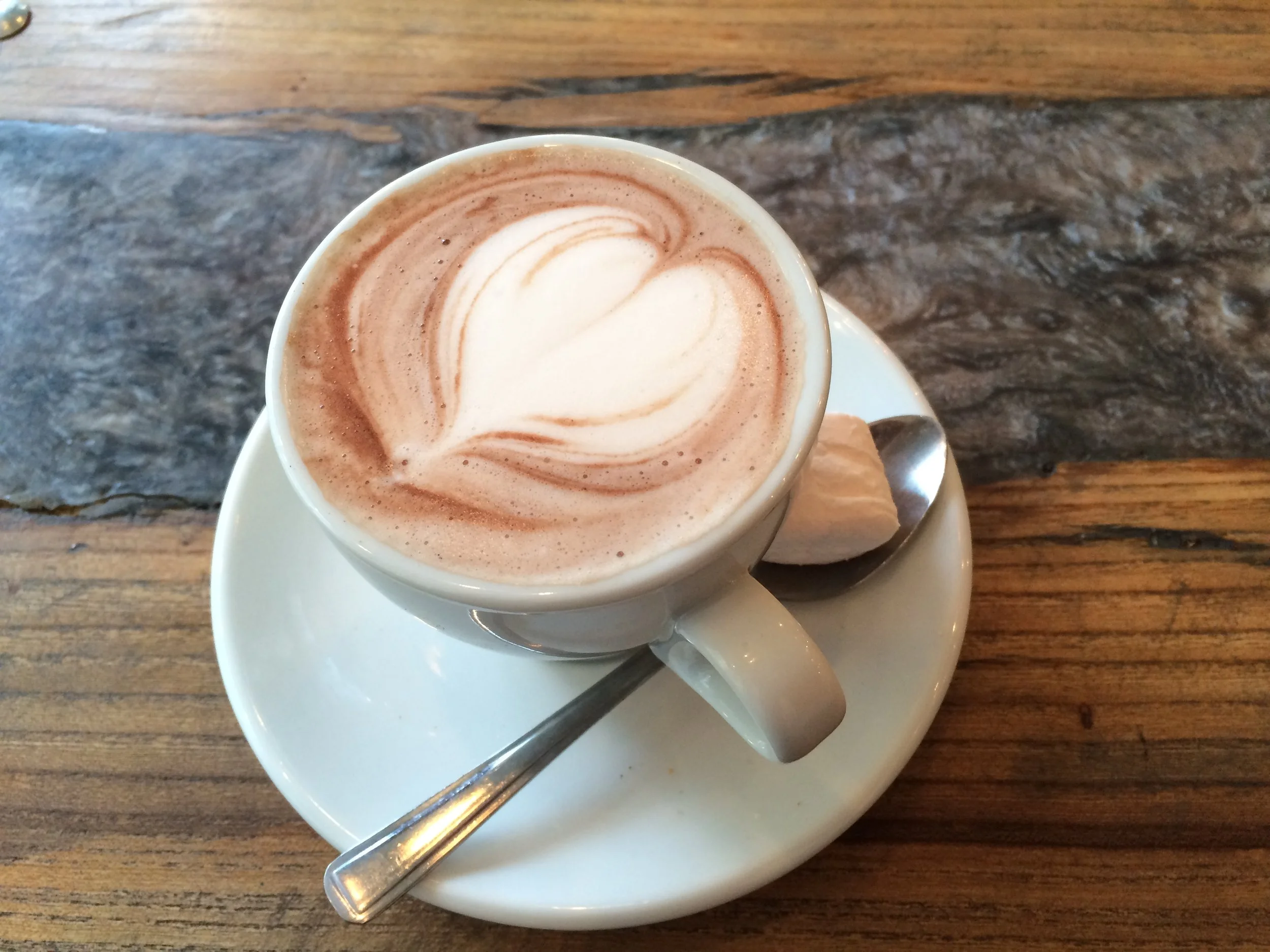 A cup of hot chocolate with whipped cream and a heart-shaped latte art design, served on a saucer with a spoon and a piece of white chocolate on a wooden table, representing mindful time.