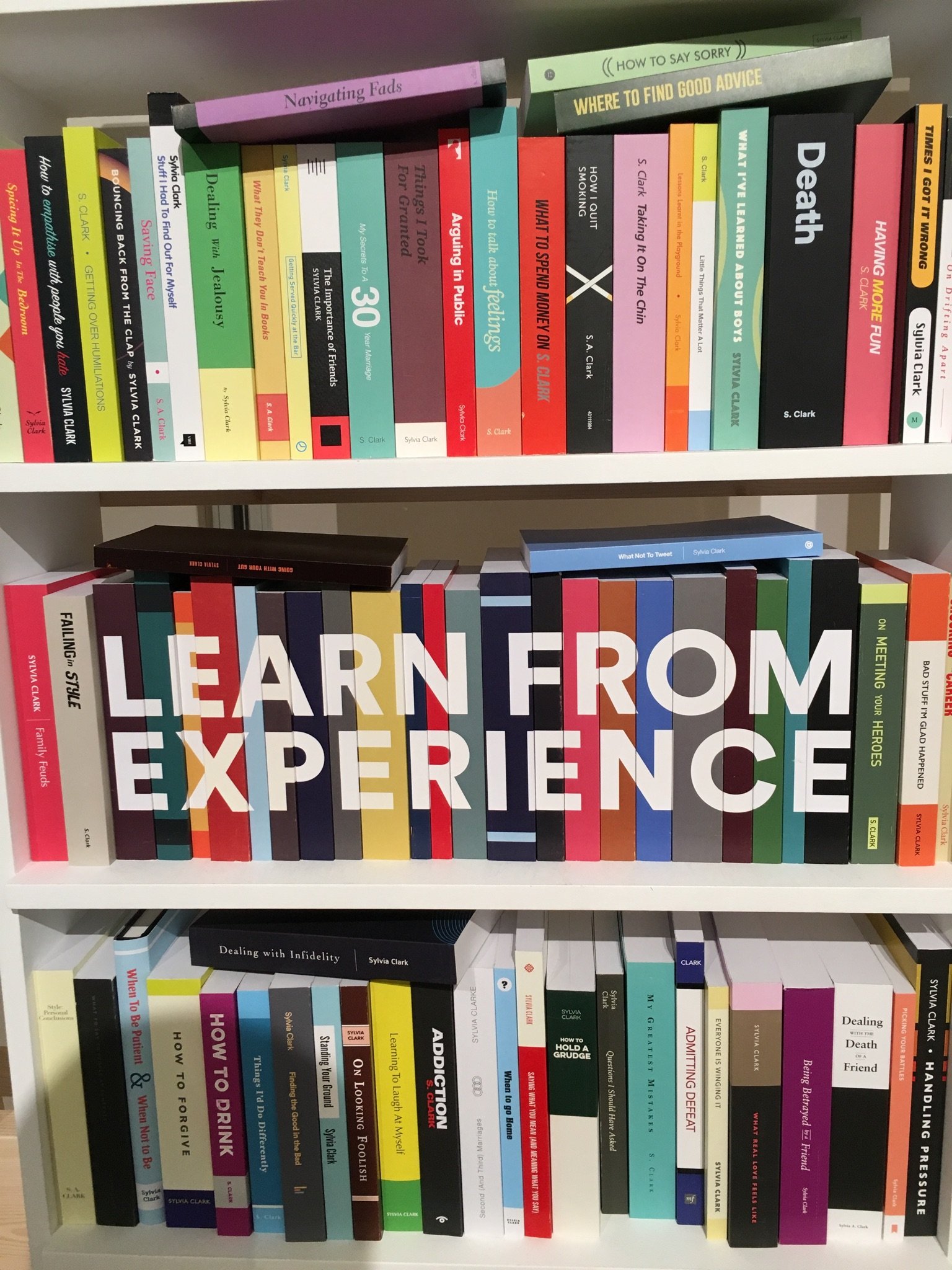 Bookshelf with colorful books organized in three rows. The middle row has books with a large sign that says 'LEARN FROM EXPERIENCE'.