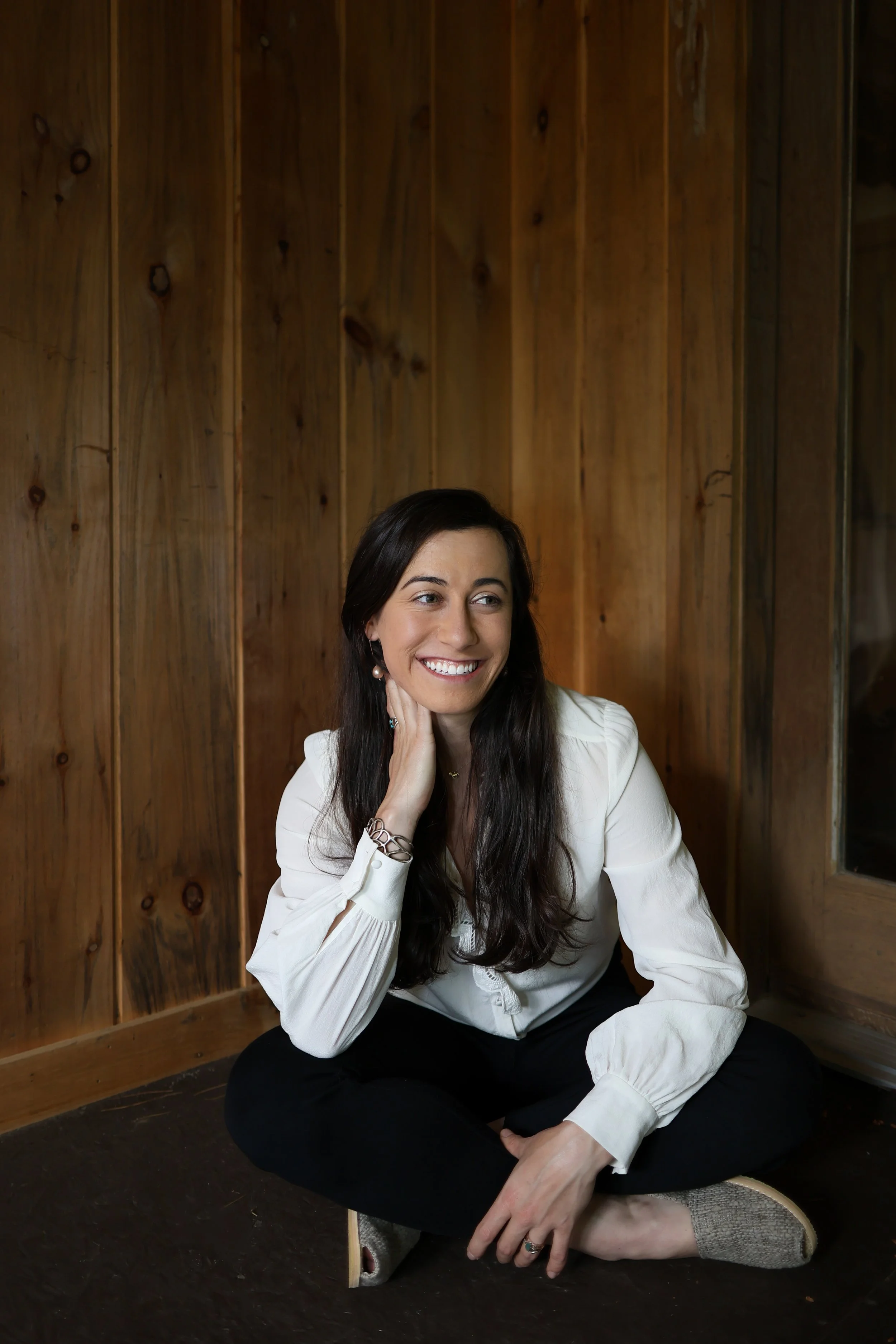 A woman sitting cross-legged on the floor, smiling and looking to her right, with long dark hair and wearing a white blouse and black pants, in a wooden room.
