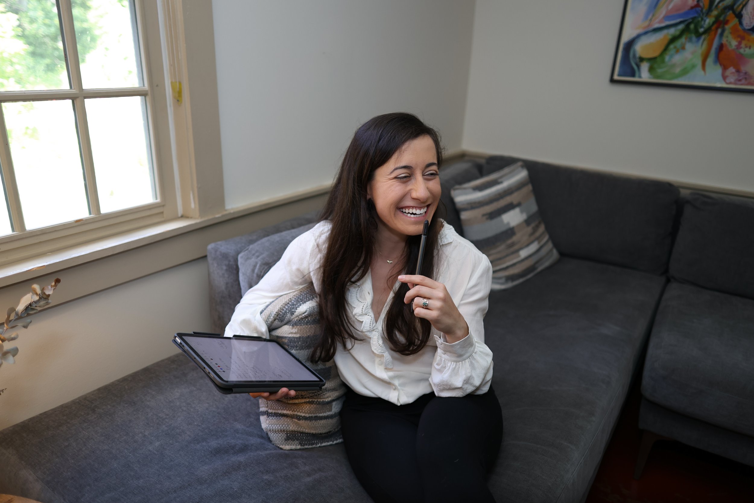 A therapist sitting on a gray sofa near a window, holding a tablet and a stylus, smiling and appearing to be in a video call or engaging in a lively conversation.