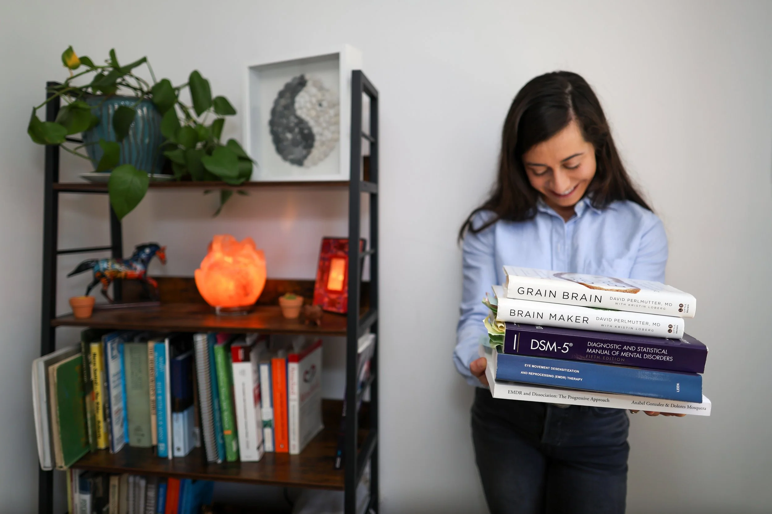 Woman holding a stack of books on mental health and psychology, standing next to a bookshelf with decorative items and books.