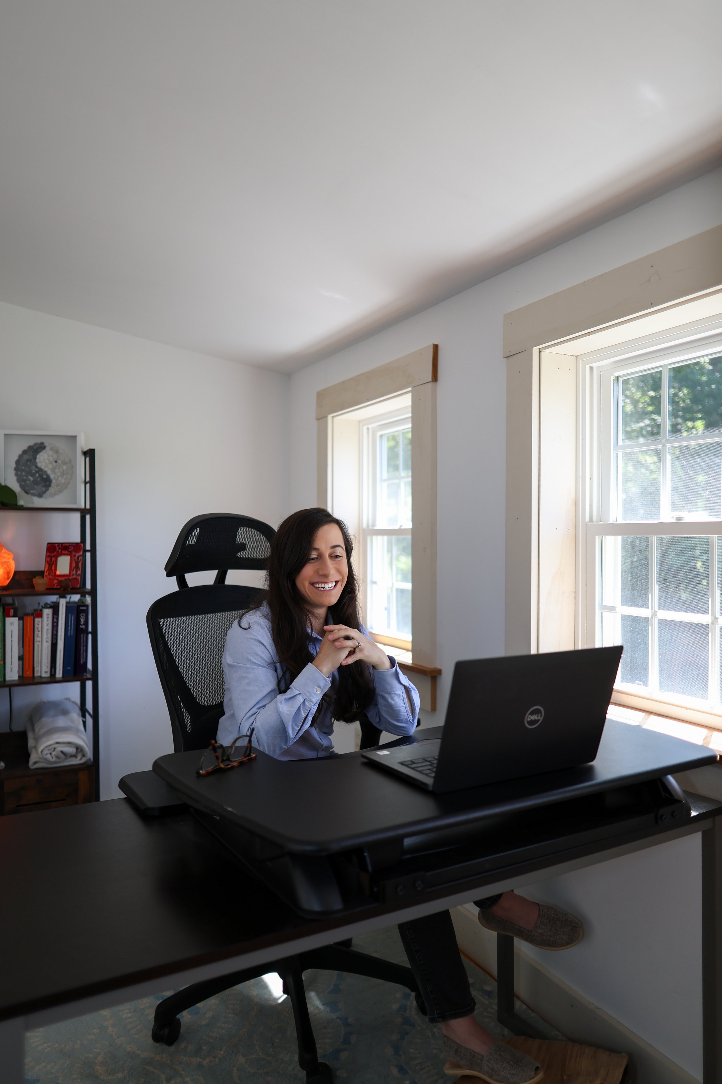A woman sitting at a desk with a Dell laptop, smiling during a video call or meeting, in a bright room with two windows letting in natural light.