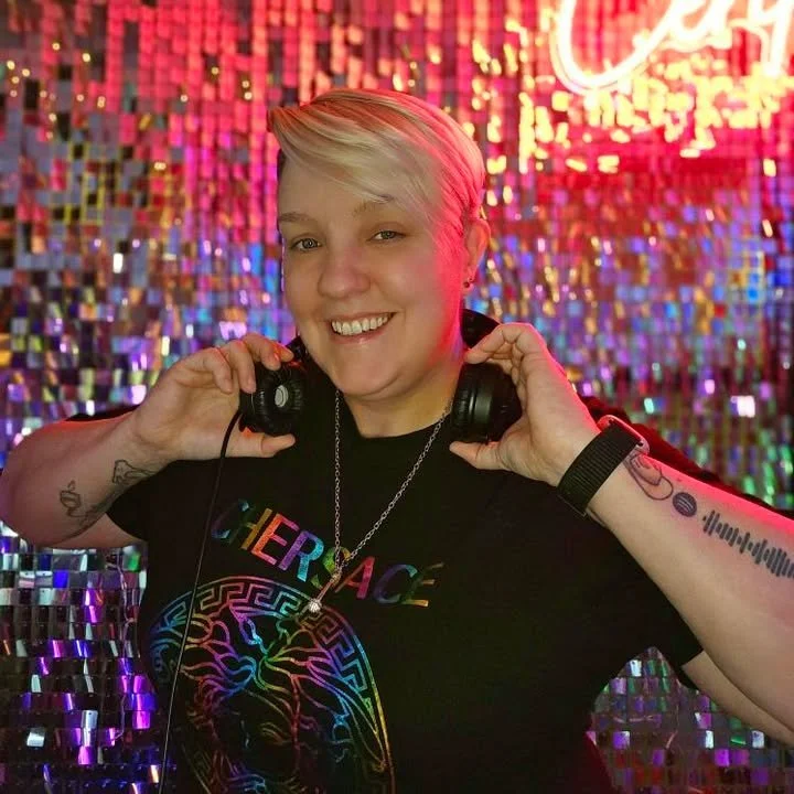 A smiling female DJ with short white hair, wearing a black top with blue straps, standing behind DJ equipment in a nightclub with green and purple lights and a crowd dancing in the background.