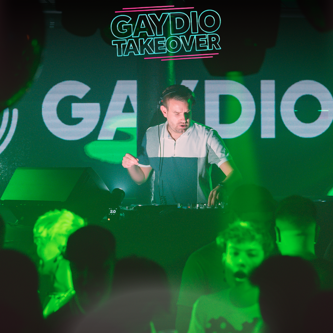 A DJ performs at an event called 'GAY DIO TAKEOVER,' with a large screen behind him displaying the text. The scene is lit with green lights, and there is a crowd of young people dancing in front of him.