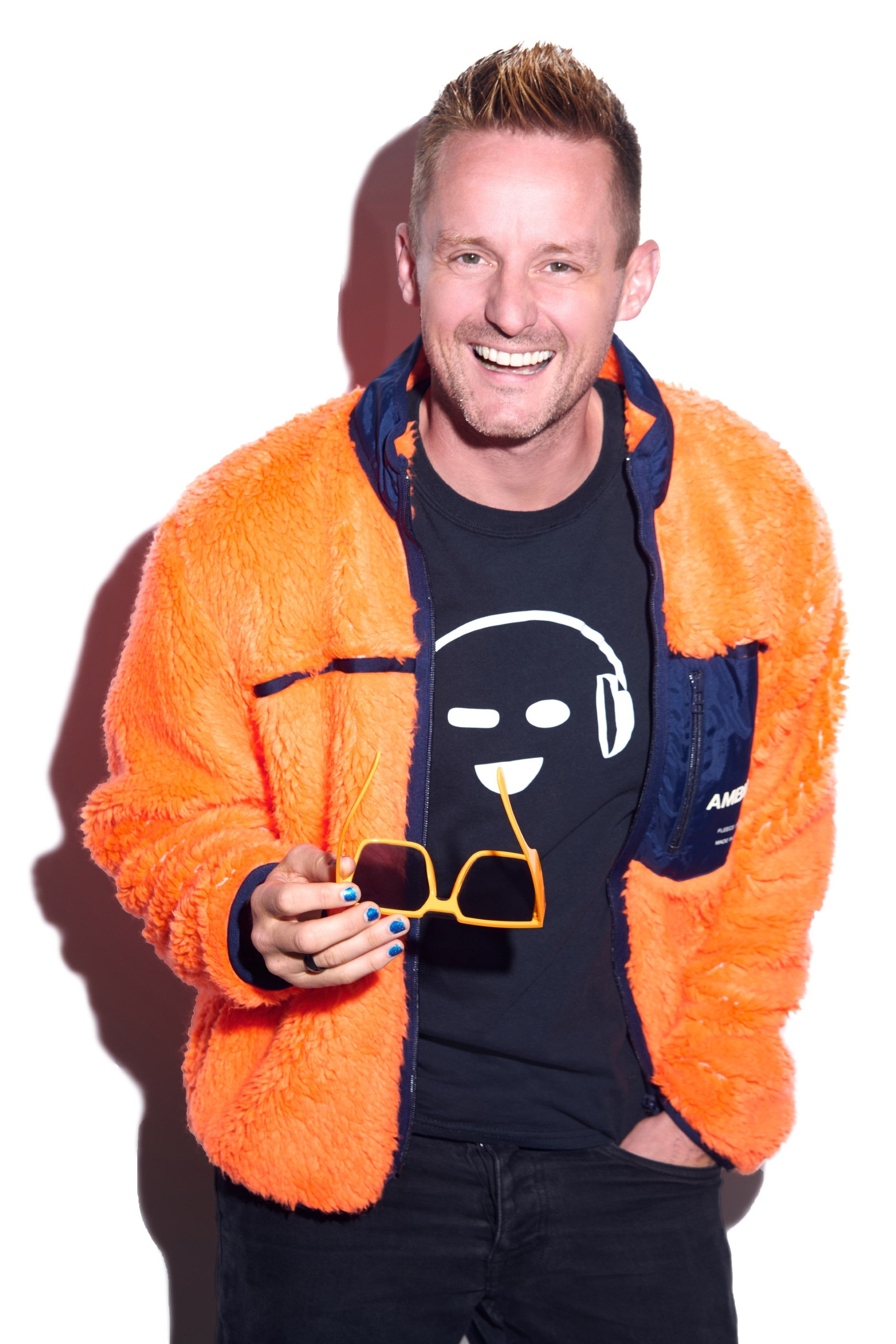 A smiling man with ginger hair and a beard wearing an orange fuzzy jacket over a black t-shirt with a headphone smiling face, holding yellow sunglasses, standing against a white background.