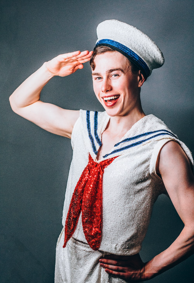 Person dressed in a sailor costume, smiling, wearing a sailor hat, salute gesture with right hand, hand on hip, sailor outfit with red sequin tie, gray background.