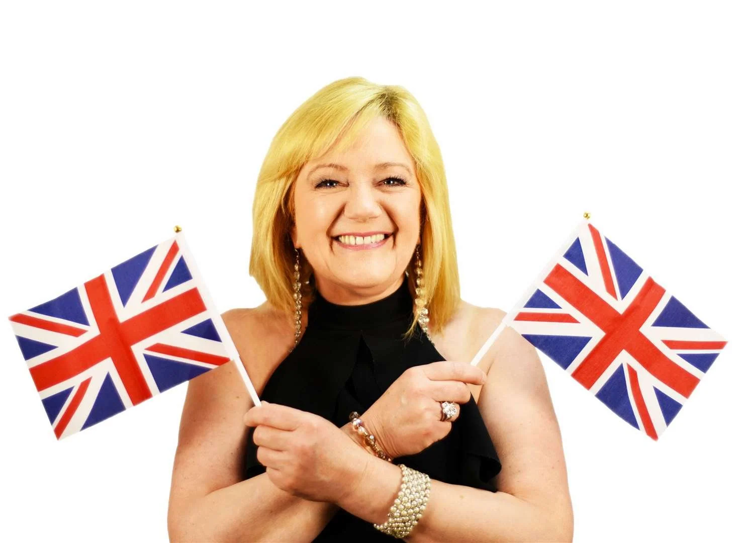A smiling woman with blonde hair holding two Union Jack flags, wearing a black sleeveless dress and jewelry, with a white background.