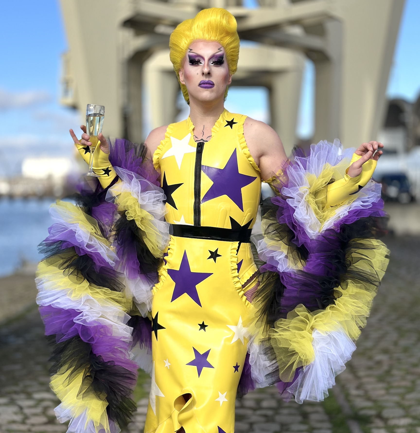 A drag queen dressed in a yellow and purple star-patterned dress with colorful tulle accessories, holding a glass of champagne outdoors near a bridge with a river and sky in the background.