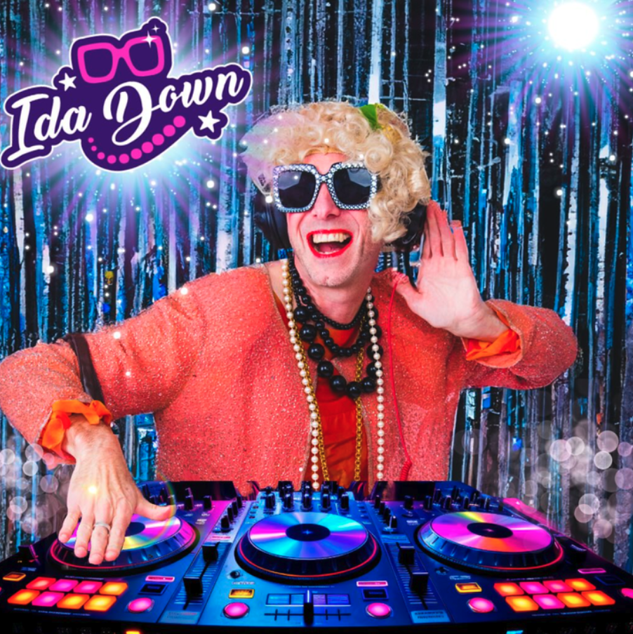 Person wearing a sparkling silver dress with red wig and feather accents and large ring, posing with arms open against a red background.