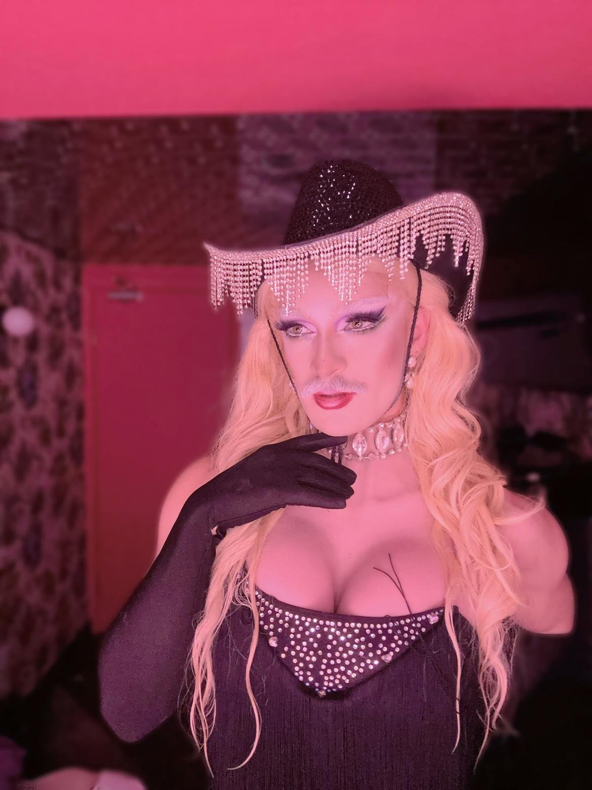 A person dressed in a black, glittery cowboy hat with a pink rhinestone fringe, black glove, and a sparkly top, posing with a contemplative expression in a pink-lit room.