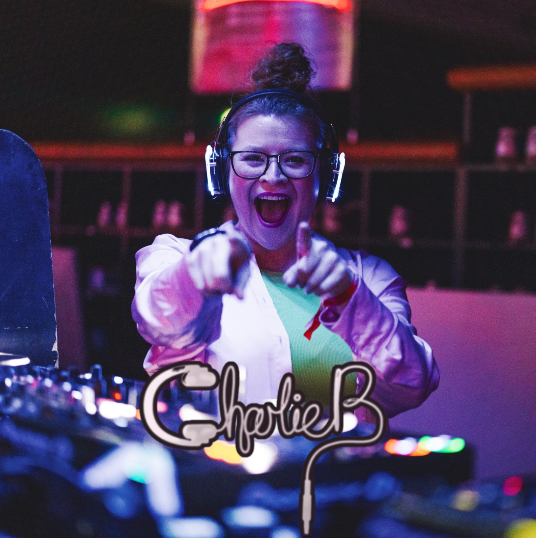 A young woman DJ with glasses, headphones, and a bun hairstyle, smiling and pointing at the camera, with a DJ mixer in front of her and colorful lights in the background.