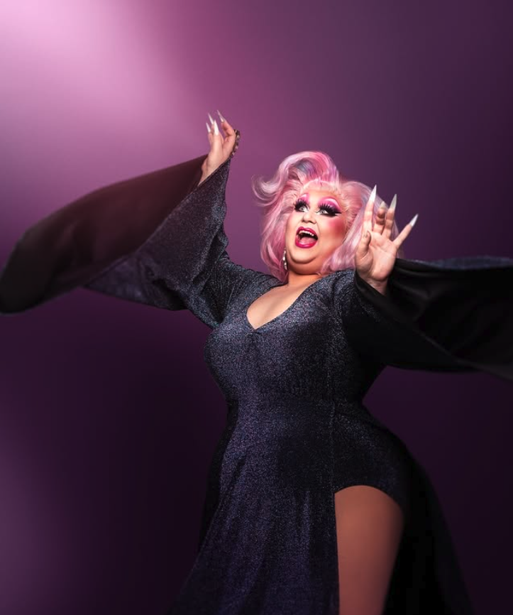 A drag queen with pink hair, dramatic makeup, and long detailed nails, wearing a sparkly black gown with a high slit, posing confidently against a purple background.