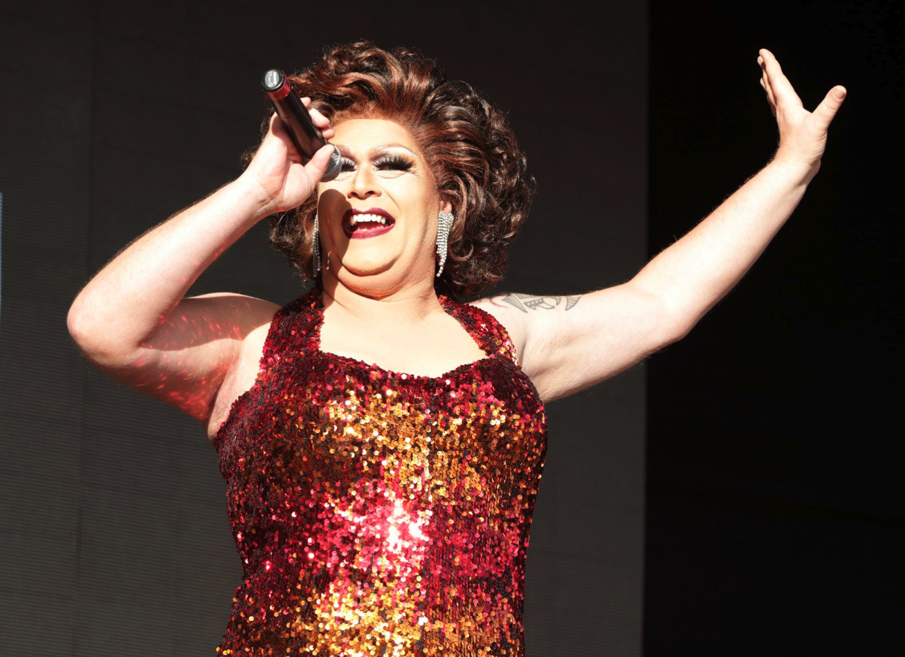 Performer singing into a microphone with one arm raised, wearing a red and gold sequined dress and large earrings.