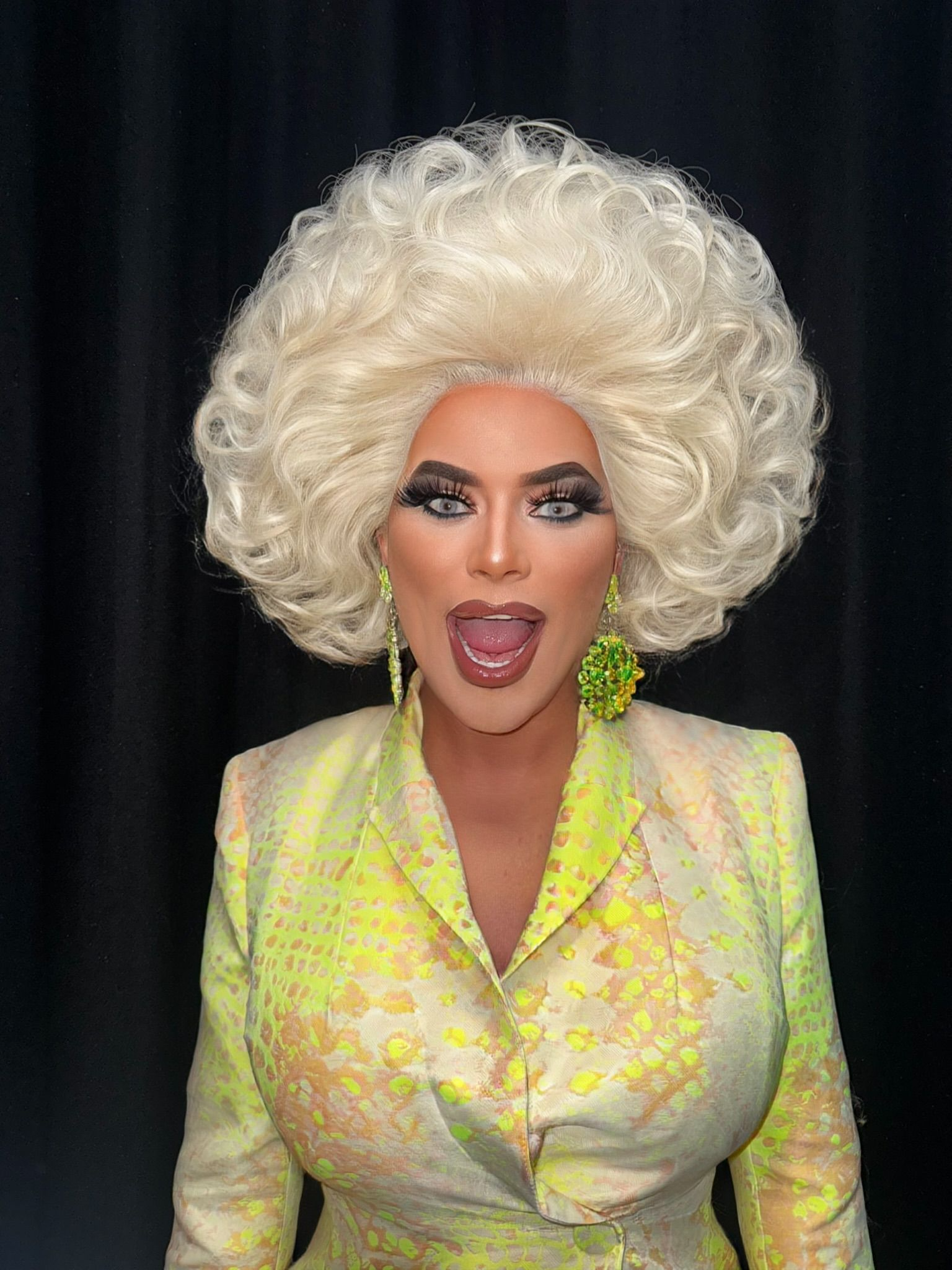 Person wearing a sparkling dress with red feather accents and large hoop earrings, smiling and posing with arms open against a gray background.