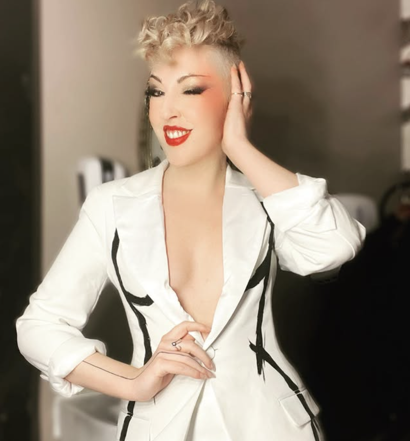 A woman with short curly blonde hair styled in a modern, edgy cut, smiling and winking. She is wearing bright red lipstick, winged eyeliner, and a white blazer with black accents, unbuttoned at the top to reveal some cleavage, and is touching her head with one hand while the other rests on her waist.