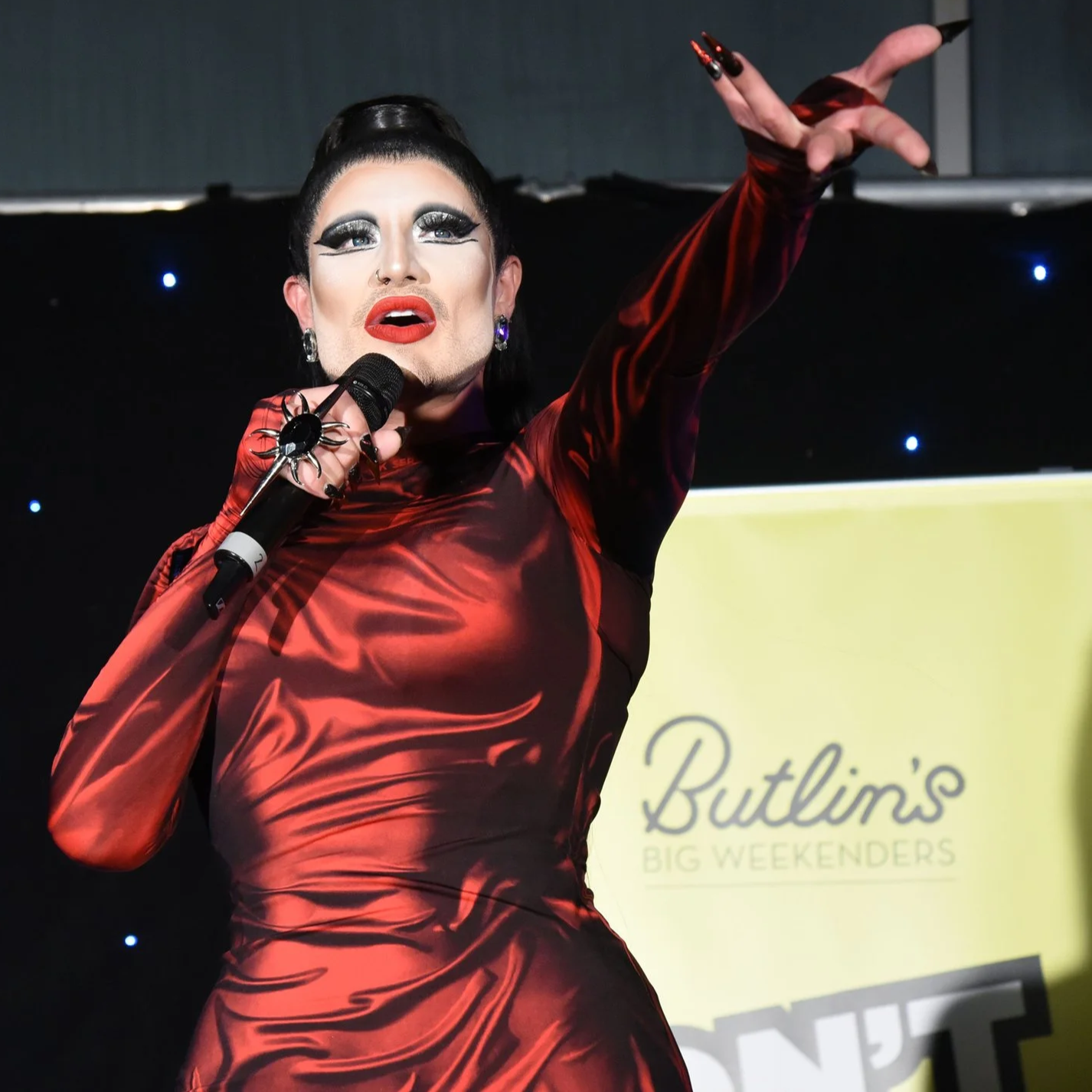 Person in black sequin suit with white collar, bold makeup, red lipstick, and elaborate black hairstyle, posing against a gray background.