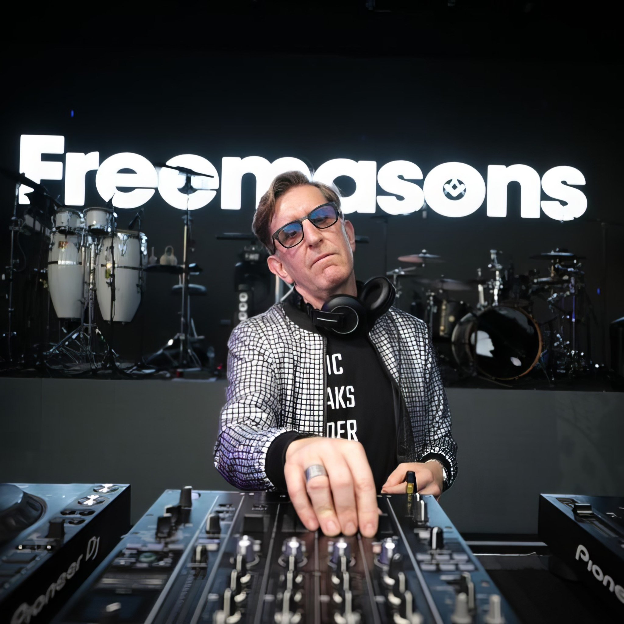 DJ at DJ deck in silver jacket black background and headphones and glasses