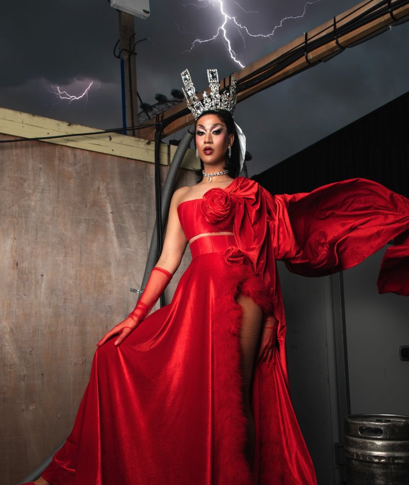 A woman in an elaborate red gown with a high slit, fishnet stocking, and dramatic cape, wearing a silver crown and jewelry, standing outdoors under a stormy sky with lightning, next to tank and pipe structures.
